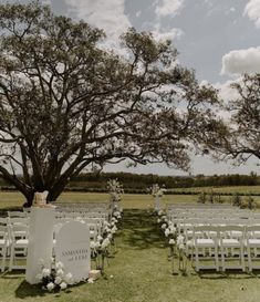 an outdoor ceremony set up with white chairs and flowers on the grass under a large tree