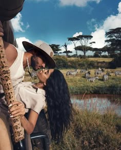 a man and woman are kissing in front of zebras on the savannah, while one is wearing a safari hat