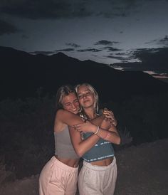 two young women hugging each other in front of a mountain at night with the sun setting behind them