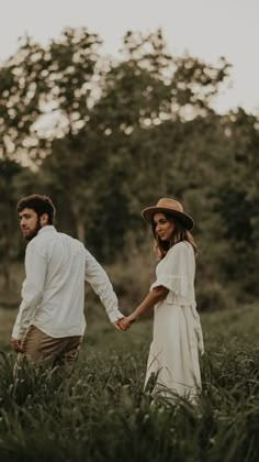 a man and woman holding hands walking through tall grass with trees in the back ground