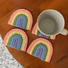 three crocheted rainbow coasters next to a coffee cup on a wooden table