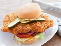 a fried chicken sandwich with pickles and tomatoes on a plate next to a napkin