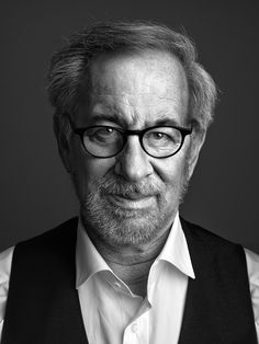 black and white photo of an older man with glasses looking at the camera while wearing a vest