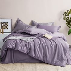 a bed with purple sheets and pillows in a room next to a potted plant