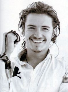 a man with long hair smiling and holding a snake in one hand while wearing a white shirt