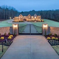 a driveway leading to a large home with lights on the front gate and landscaping around it