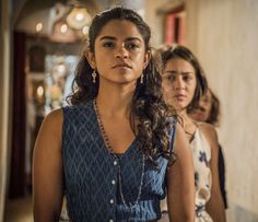 two women standing in a hallway one is wearing a blue top and the other wears a white dress