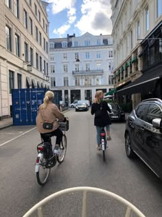 two women riding bikes down the middle of a street with cars parked on both sides