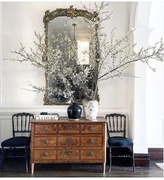 a room with a dresser, mirror and vase on the wall next to two chairs