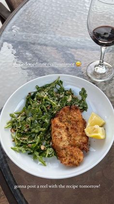 a white plate topped with meat and greens next to a glass of wine