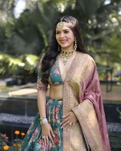 a woman in a green and pink lehenga standing next to some flowers with her hands on her hips