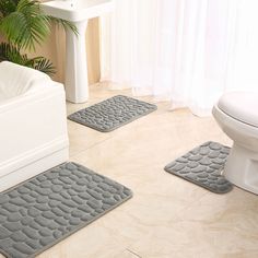 three bathroom rugs on the floor in front of a toilet and bathtub with plants next to it