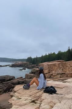 a woman sitting on top of a rocky cliff next to the ocean with a backpack