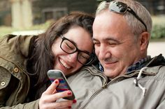 an older man and young woman are smiling while looking at a cell phone together in the street