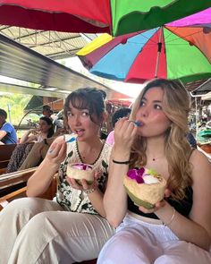 two women sitting under an umbrella eating sandwiches and drinking juice from their cups in front of them