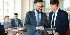 two men in suits look at something on a clipboard while other people are working behind them