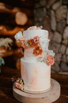 a white and pink wedding cake with flowers on top sitting on a wooden table in front of a stone wall