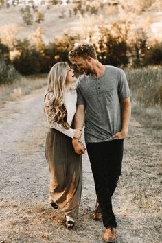 a man and woman walking down a dirt road in front of some trees with their hands around each other