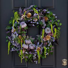 a wreath with purple flowers and green leaves hangs on a black front door, surrounded by greenery