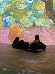 two people are laying on the floor in front of a colorful wall with trees and clouds