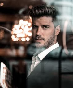 a man with a goatee and suit looks out the window in front of a chandelier