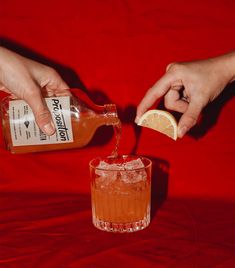 two hands are pouring orange juice into a glass with an orange slice on the rim