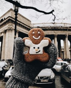 a person holding up a ginger cookie in front of a building with cars parked on the street