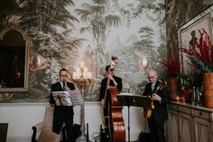 two men are playing instruments in front of a wall with trees and plants on it