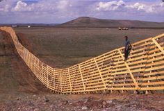 a man standing on top of a wooden fence in the middle of nowhere with mountains in the background