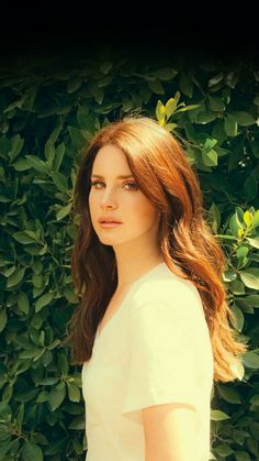 a woman with long hair standing in front of some bushes and looking at the camera