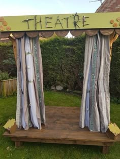 an outdoor stage with curtains and the words theatre written on it's sides, in front of a garden