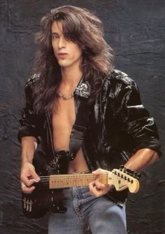 a man with long hair is holding a guitar and posing in front of a black background