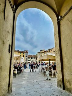 people are walking through an archway in the middle of a town square with tables and umbrellas