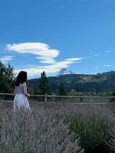 a woman in a white dress standing next to some lavender bushes and trees with mountains in the background