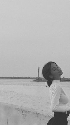 black and white photograph of a woman standing by the water looking up at a kite flying in the sky