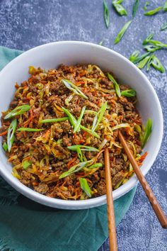 a white bowl filled with meat and vegetables next to chopsticks on a blue cloth