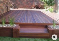 a wooden deck surrounded by plants and rocks on the grass in front of a brick building