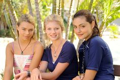 three young women sitting on a bench in front of palm trees and looking at the camera