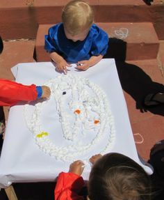 two small children are sitting at a table making something out of white string and plastic beads