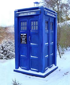 a blue phone booth sitting in the middle of a snow covered field with trees and bushes behind it