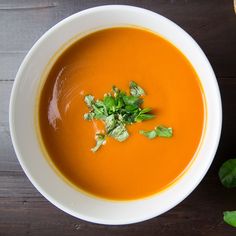 a bowl of carrot soup next to some bread on a wooden table with fresh herbs