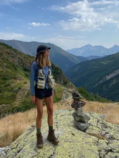 a woman standing on top of a mountain next to a pile of rocks and grass