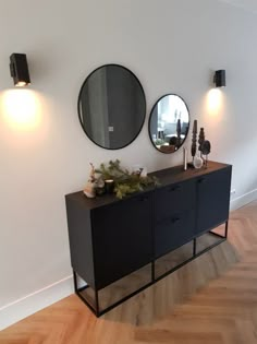 two round mirrors on the wall above a black buffet table with plants and vases