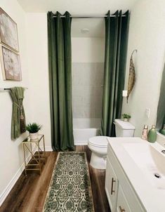 a white bathroom with green curtains and rugs on the wooden floor next to the toilet