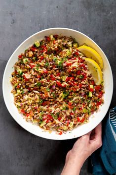 a person holding a white bowl filled with rice and veggies next to lemon wedges