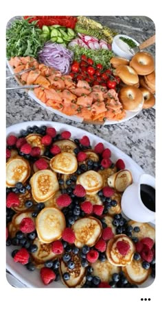 two plates filled with pancakes and berries next to a platter full of fruit on a table