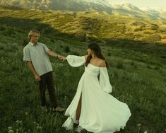 a man and woman holding hands while standing in the middle of a field with mountains behind them