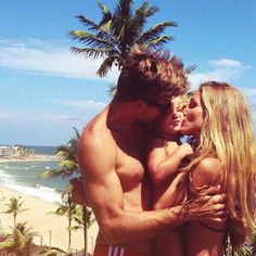 a man and woman embracing each other in front of the ocean with palm trees behind them