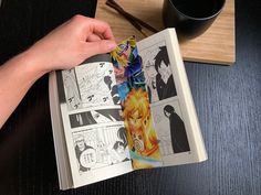 a person is holding an open comic book on a table with a cup of coffee