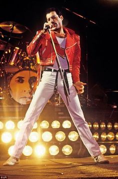 a man in red jacket and white pants on stage holding a microphone with lights behind him